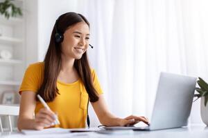 Smiling young korean woman using headset while working at home office desk photo