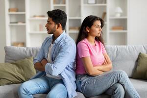 Unhappy indian couple sitting apart on sofa with arms crossed photo