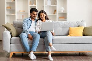 Relaxed indian spouses resting on couch with laptop smiling photo