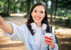 Delighted young woman with headphones taking a selfie and holding a red coffee cup photo