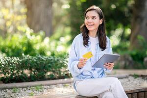 Radiant young woman sitting on a park bench holds a golden credit card and tablet photo