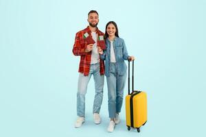 Cheerful caucasian travelers couple standing with tickets and suitcase, studio photo