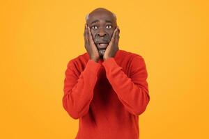 Shocked senior black man with hands on cheeks and wide eyes, wearing a red sweater photo