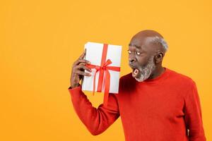 Shocked senior black man in a red sweater holding a white gift box with a red ribbon photo