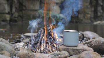A mug on a stone near a fire on the river bank, close-up. Travel concept. 4k video