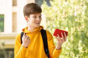 emocionado contento adolescente chico ganador sensación alegría sonriente y mirando a teléfono inteligente chico utilizando teléfono, victorioso lotería juego, consiguiendo devolución de dinero, en línea regalo en aplicación, participación célula teléfono exterior. foto