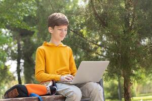 Smiling teenager boy working on laptop. Holding and using a laptop for networking on a sunny spring day, outdoors. photo