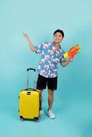 Full body photo of handsome Asian tourist in summer clothes with water gun and luggage during Songkran festival on blue background with copy space.