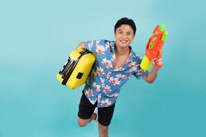 Handsome Asian tourist in summer clothes with water gun and luggage during Songkran festival on blue background. photo
