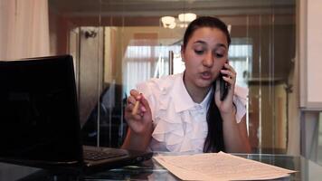 Beautiful young business girl with dark hair in business clothes talking on a mobile phone discussing a business project in documents in the office sitting at a table. HD video