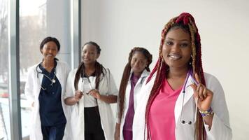 A young african american doctor with red hair stands in front and looks at the camera against the background of a team of colleagues video