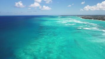 dar visie van blauw caraïben zee met koraal rif en kustlijn met wit zand strand en huizen omringd door groen tropisch Woud met bewolkt lucht Aan een zonnig dag video