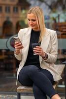 woman in a white jacket and black pants is sitting on a bench with a cell phone in her hand and a cup of coffee photo