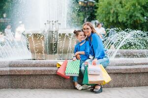 Beautiful mom and her cute little daughter are holding shopping bags, looking at camera and smiling while standing outdoors. Shopping concept photo