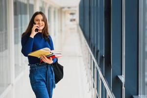 Cheerful brunette student girl with black backpack holds books in modern building. female student standing with books in college hallway photo