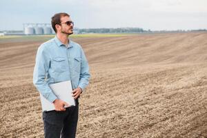 cosecha concepto. granjero en un campo con un ordenador portátil en un antecedentes de un agrícola silos para almacenamiento y el secado de granos, trigo foto