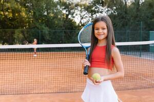 niño jugando tenis en al aire libre corte. pequeño niña con tenis raqueta y pelota en deporte club. activo ejercicio para niños. verano ocupaciones para niños. formación para joven niño. niño aprendizaje a jugar foto