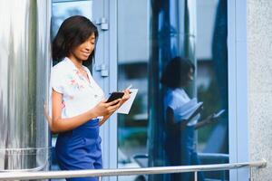 View from below - cute African American girl typing text on smart phone standing in the middle of big business center in the city on summer day photo