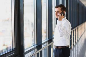 retrato de un grave confidente hombre jefe teniendo móvil teléfono conversacion mientras descansando después reunión con su socios, empresario hablando en célula teléfono mientras en pie en moderno espacio adentro foto