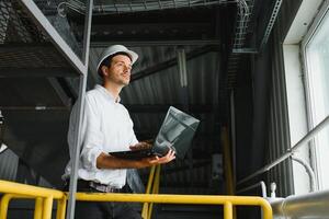 A young handsome employee of a modern factory in a white hard. In the background is a large shop for the production. photo
