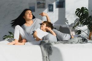 Young mother with her 5 years old little son dressed in pajamas are relaxing and playing in the bed at the weekend together, lazy morning, warm and cozy scene. Family home shoot. photo