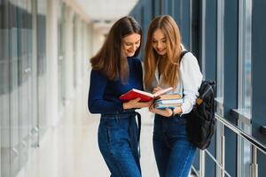 Two young female students standing with books and bags in the hallway University speaking each other. photo