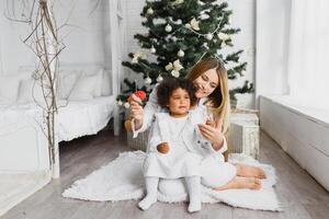 Merry Christmas and Happy Holidays Cheerful mom and her cute daughter at Christmas tree. Parent and little child having fun near Christmas tree indoors. Loving family with presents in room. photo