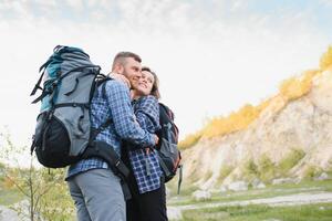 Pareja de joven contento viajeros excursionismo con mochilas en el hermosa rocoso sendero a calentar soleado noche. familia viaje y aventuras concepto. foto
