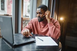 afro americano negociaciones vídeo charla en computadora se sienta en café con taza de café. negro hombre llamadas en ordenador portátil en vídeo enlace y habla él usa en camisa y traje chaqueta. vídeo enlace en línea conexión foto