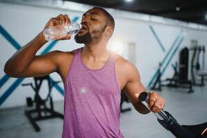 young african american sportsman drinking water and looking away in gym. photo
