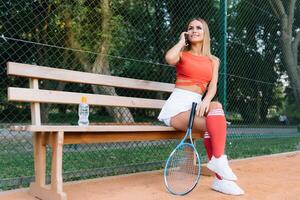 Portrait of fashionable woman in red and white clothing with tennis racket posing at tennis net on court. Sports Fashion photo