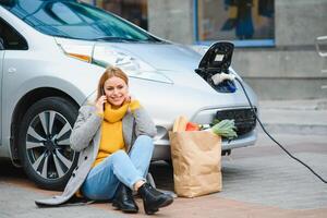 usando el teléfono inteligente mientras espera. mujer en la estación de carga de coches eléctricos durante el día. vehículo nuevo foto