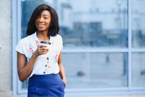 hermosa africano americano niña en pie en calle con Teléfono móvil y café en manos mientras felizmente mirando aparte. retrato de africano americano dama en calle. foto