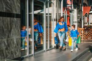 Beautiful mom and her cute little daughter are holding shopping bags, looking at camera and smiling while standing outdoors. Shopping concept. photo