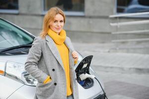 Just wait. Pleasant cute blonde woman leaning on her car charging it and looking aside photo