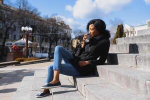 pensativo africano mujer vistiendo lentes Bebiendo tomar lejos café sentado en el calle escalera foto