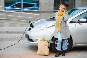 tener una conversación usando el teléfono. mujer en la estación de carga de coches eléctricos durante el día. vehículo nuevo foto