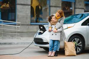 madre con hija cargando electro coche a el eléctrico gas estación y hablar en móvil teléfono foto