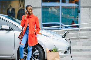 utilizando teléfono inteligente mientras espera. africano americano niña en el eléctrico carros cargar estación a tiempo de día. marca nuevo vehículo foto