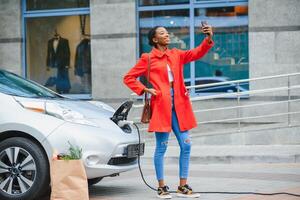 cargando electro coche a el eléctrico gas estación. mujer en pie por el coche. dama con productos alimenticios foto