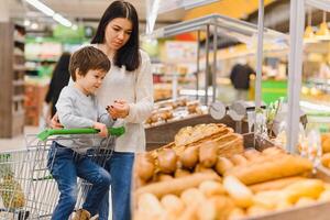 Cute child with mother choosing baked goods or pastry in bakery shop. Little boy with mom buying bread or buns at grocery supermarket photo