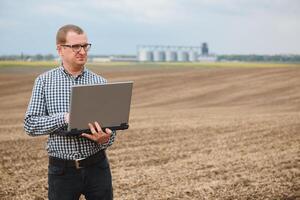 cosecha concepto. granjero en un campo con un ordenador portátil en un antecedentes de un agrícola silos para almacenamiento y el secado de granos, trigo foto