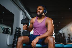 joven africano americano hombre sentado y levantamiento un pesa cerca a el estante a gimnasia. masculino peso formación persona haciendo un bíceps rizo en aptitud centro. foto