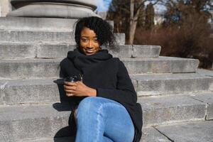 alegre oscuro desollado mujer disfrutando café participación a Vamos taza recreando en ciudad parque, contento de moda vestido africano americano hipster niña sentado al aire libre durante soleado día Bebiendo bebida foto