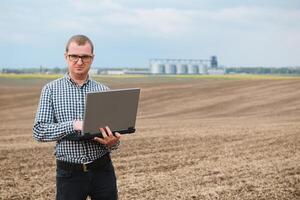 cosecha concepto. granjero en un campo con un ordenador portátil en un antecedentes de un agrícola silos para almacenamiento y el secado de granos, trigo foto