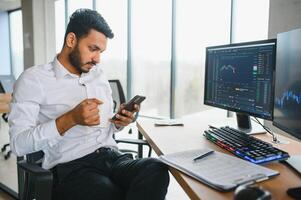 Side view of smart Indian crypto trader, investor, analyst broker, using laptop and smartphone analyzing digital cryptocurrency exchange, stock market charts, thinking of investing and funds risks photo