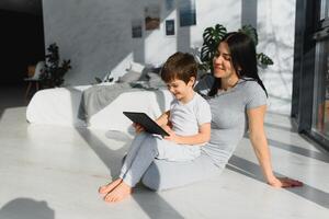 linda pequeño chico y su joven madre utilizando un digital tableta y sonriente en el piso a hogar. contento familia con artilugio foto