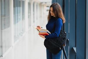 alegre morena estudiante niña con negro mochila sostiene libros en moderno edificio. hembra estudiante en pie con libros en Universidad pasillo foto