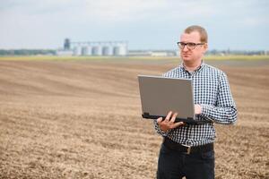 moderno granjero comprobación su campo planta y trabajando en ordenador portátil computadora en contra maíz secadora silos en concepto de industrial y agricultura foto