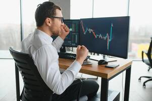 Man trader in formalwear sitting at desk in frot of monitors with charts and data at office browsing laptop checking documents analyzing stocks price changes concentrated. photo